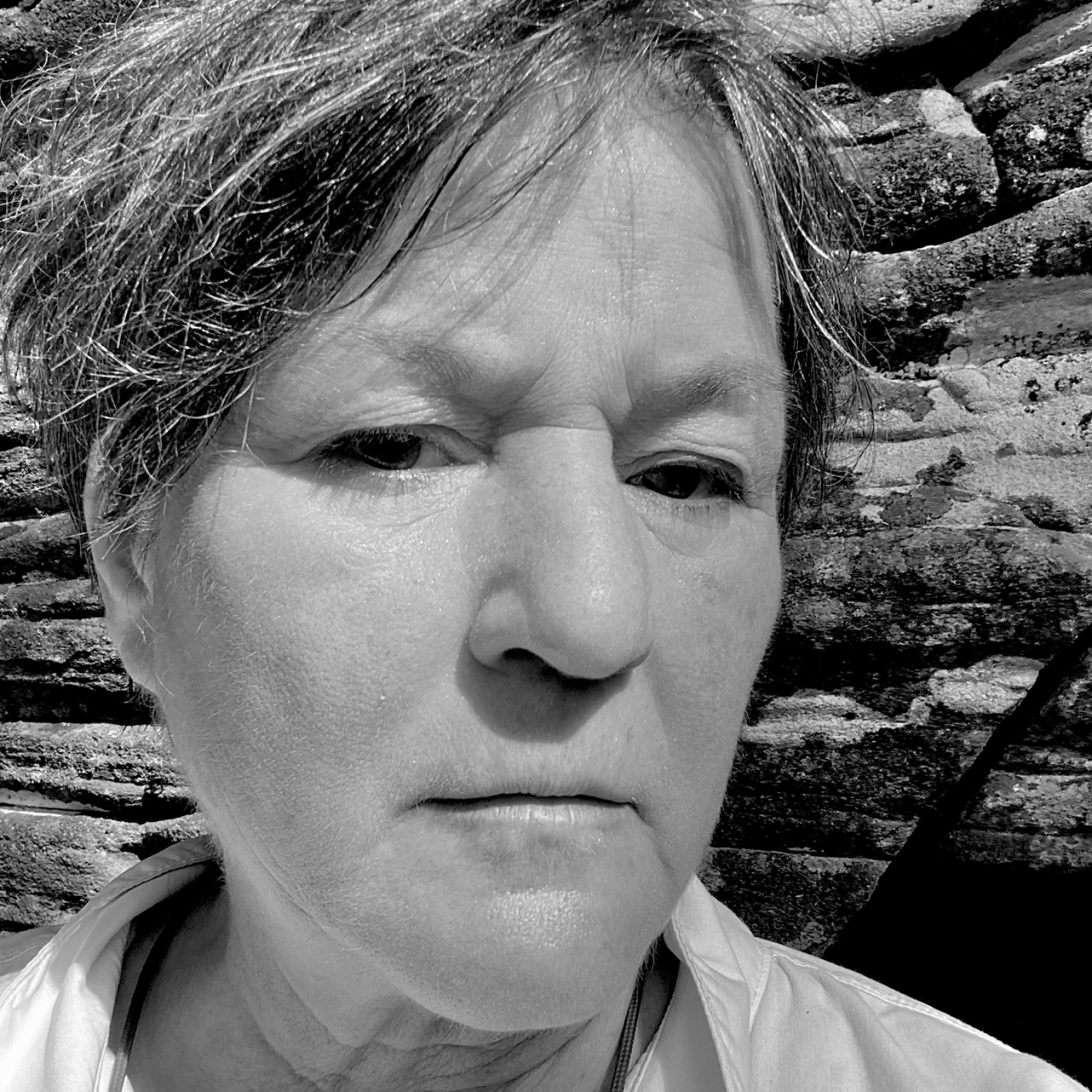 A grayscale close-up portrait of a woman with short, windblown hair standing outdoors. The woman has a neutral expression on her face, and textured rock formations are visible in the background.