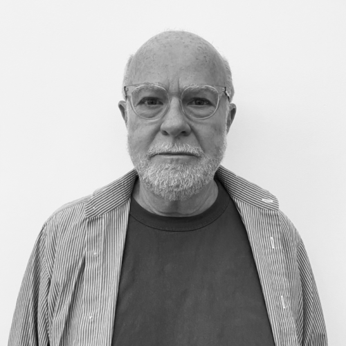 A black and white portrait of an older man with glasses and a short beard, wearing a striped shirt over a dark t-shirt, standing in front of a white wall with a neutral expression.