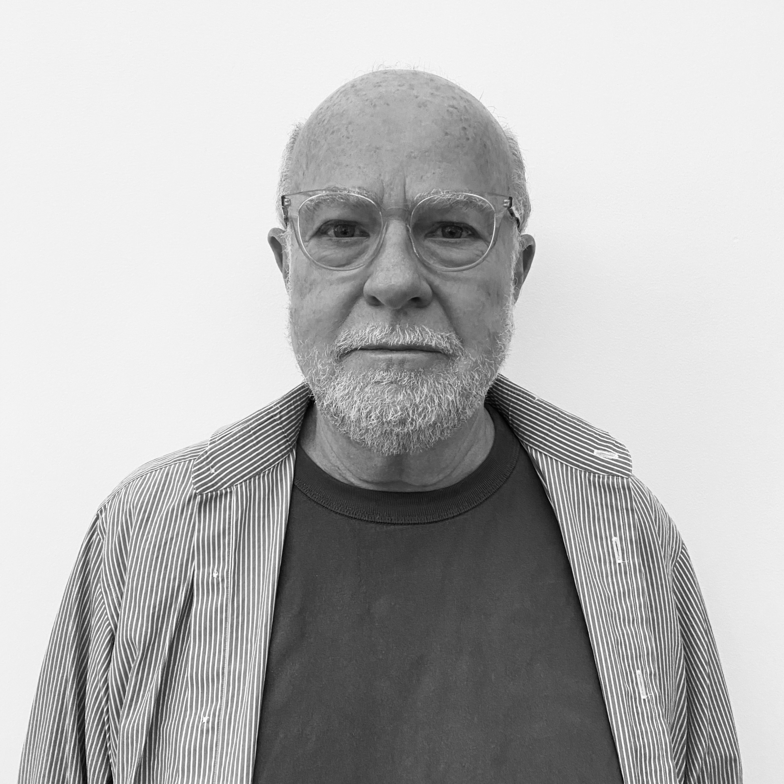 A black and white portrait of an older man with glasses and a short beard, wearing a striped shirt over a dark t-shirt, standing in front of a white wall with a neutral expression.