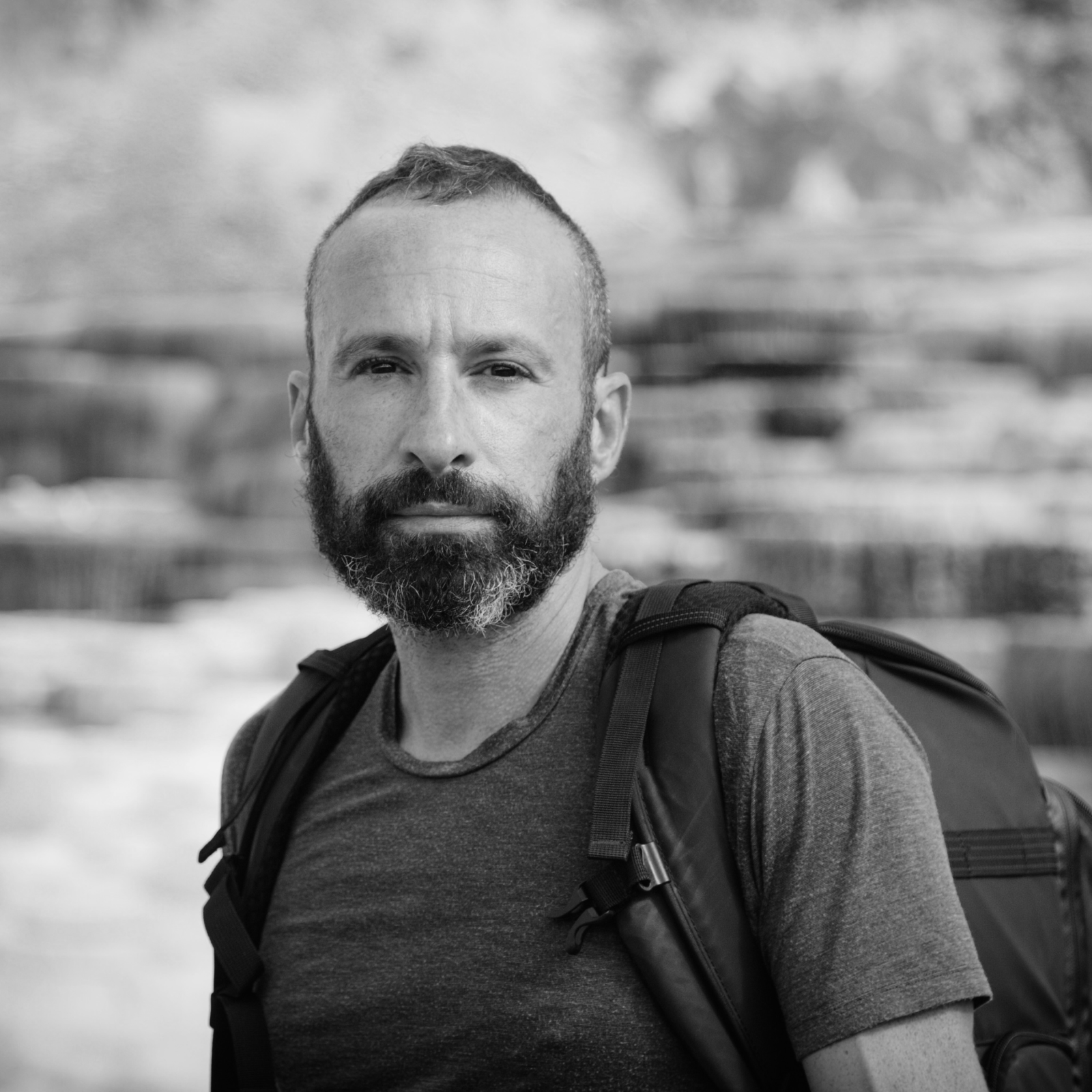 A black and white portrait of a man with a beard, wearing a t-shirt and a backpack, standing in an outdoor setting with blurred trees and rocks in the background.