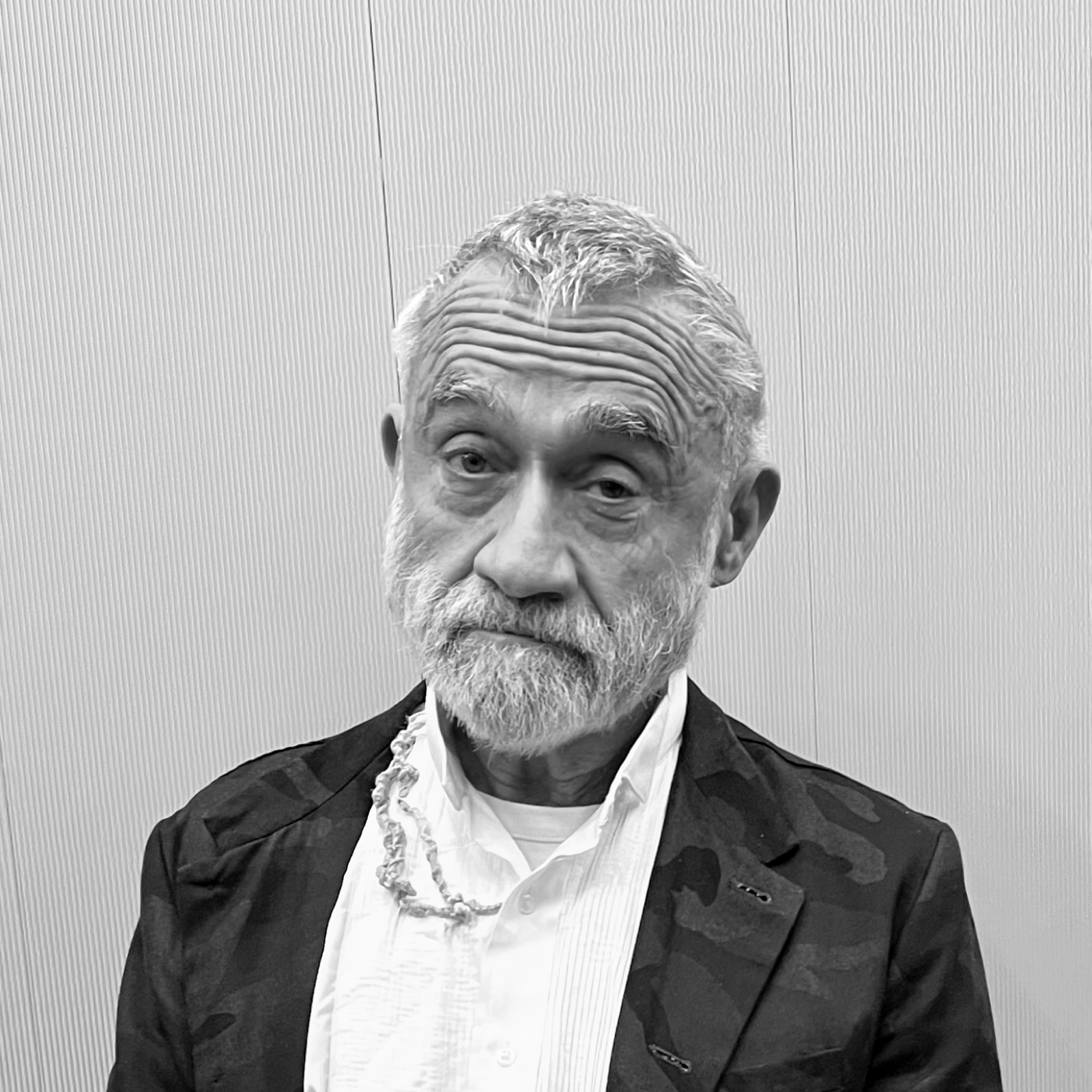 A grayscale portrait of an older man with a neatly trimmed beard and short hair. He is wearing a suit jacket over a collared shirt and has a thoughtful expression, standing against a plain background.