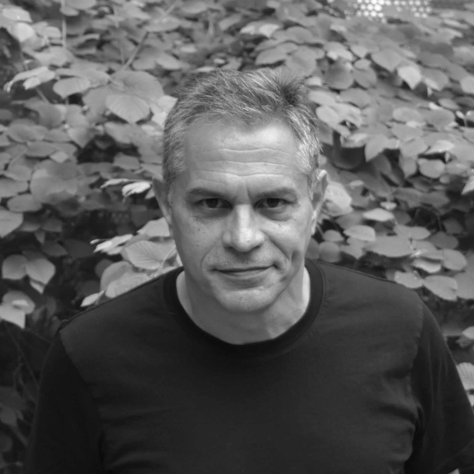 A black-and-white portrait of Ignacio Urbina Polo, standing in front of trees, wearing a dark shirt, smiling at the camera.