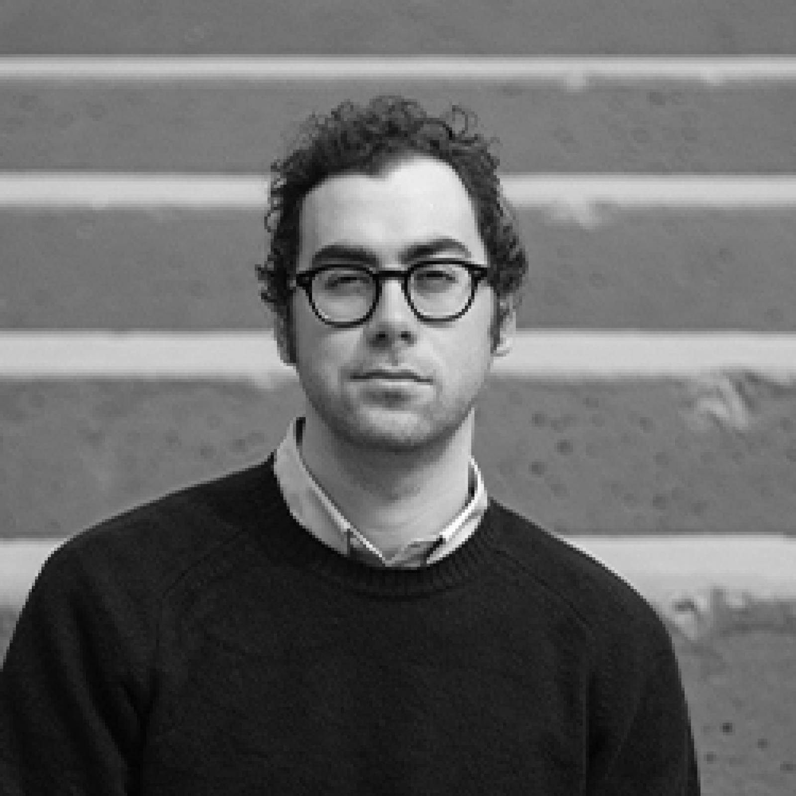 A grayscale portrait of Jacob Turetsky, a man with curly hair and round glasses, standing on a staircase, wearing a sweater and looking directly at the camera.