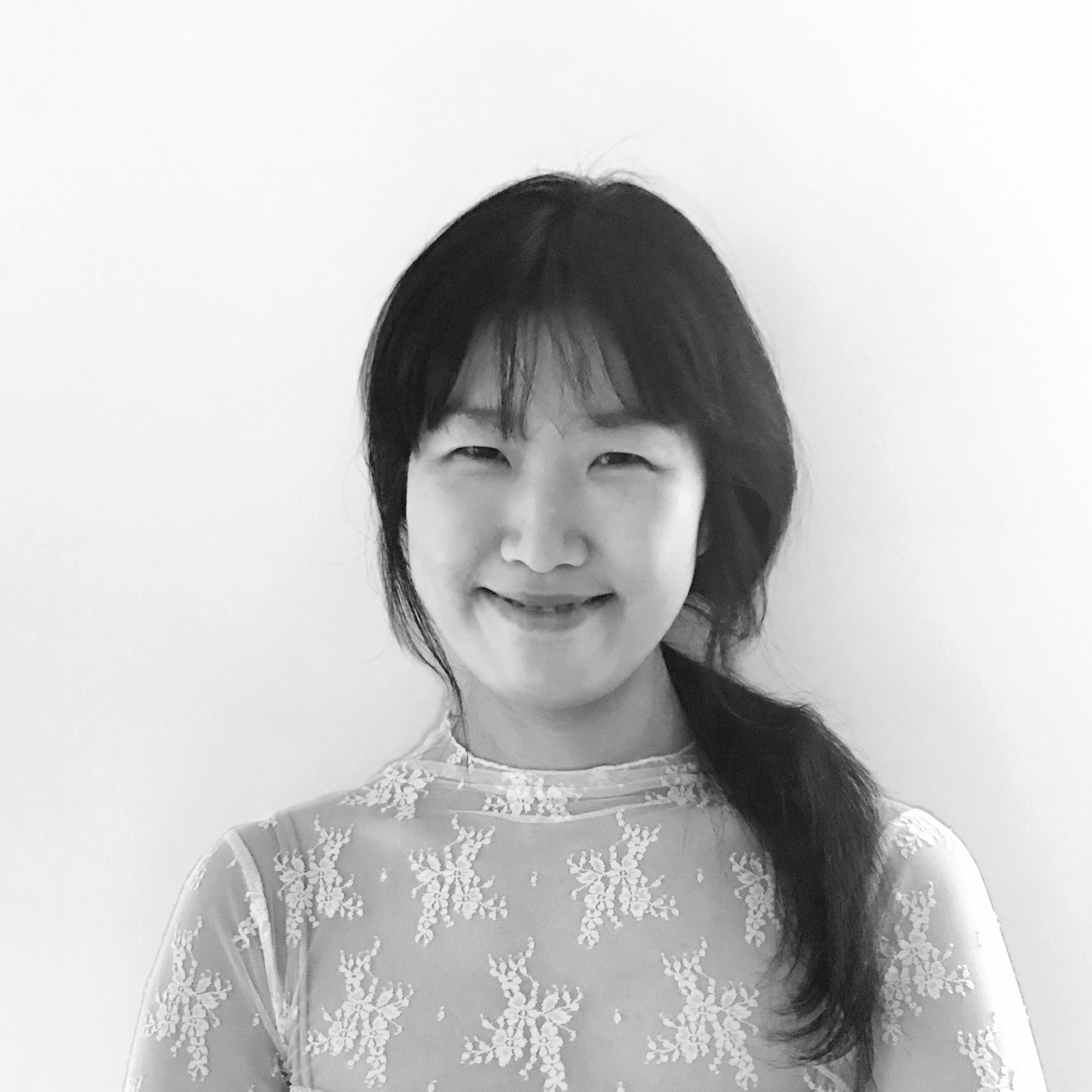 A black and white portrait of a woman with a side ponytail, wearing a lace top and smiling softly at the camera.