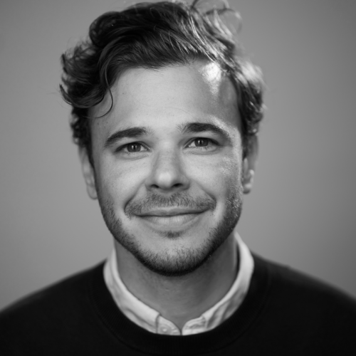 A black and white portrait of a man with curly hair and a slight beard, wearing a white shirt under a sweater, smiling at the camera.