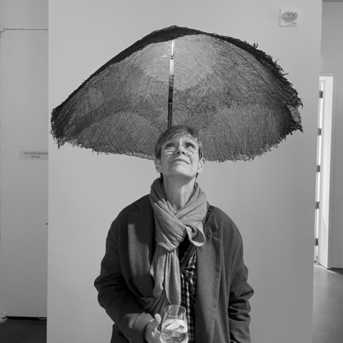 Kate Hixon smiles slightly while standing under an art installation resembling an umbrella, indoors at an exhibition.