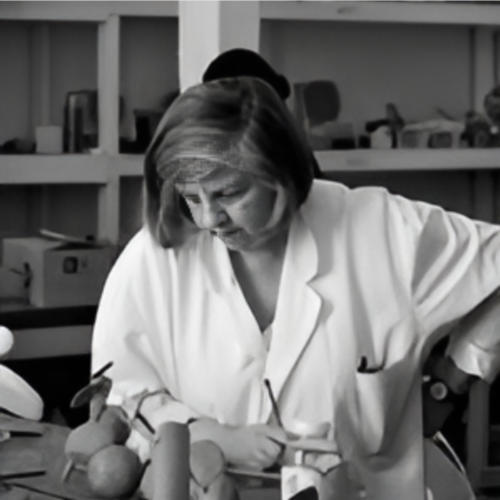 A grayscale image of a woman wearing a lab coat, focusing intently on her work at a table filled with various objects and tools. She appears to be carefully handling one of the objects.