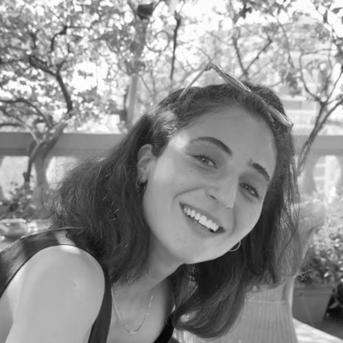 A black and white portrait of a woman with shoulder-length hair, smiling in an outdoor setting, with trees and sunlight in the background.