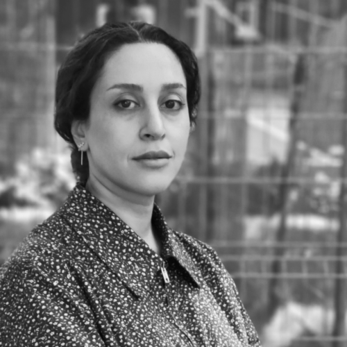 A grayscale portrait of Mahtab Pedrami, a woman with her hair pulled back, wearing a patterned jacket, standing outdoors in front of a blurred background of fencing and greenery.