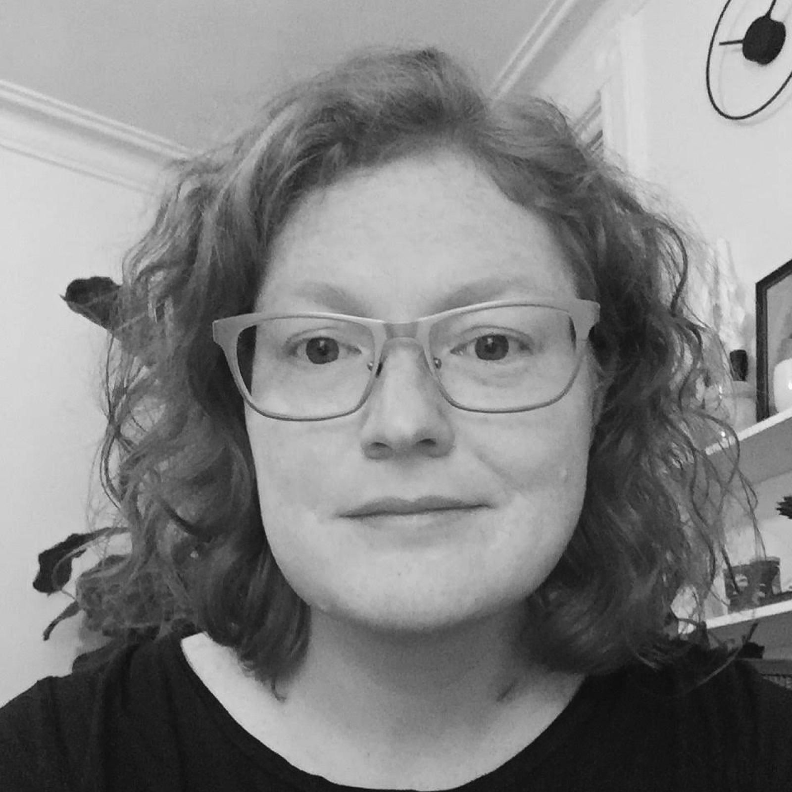 A grayscale close-up portrait of a woman with curly, shoulder-length hair, wearing glasses and a black top. She has a neutral expression and is sitting indoors, with a bookshelf and a plant visible in the background.