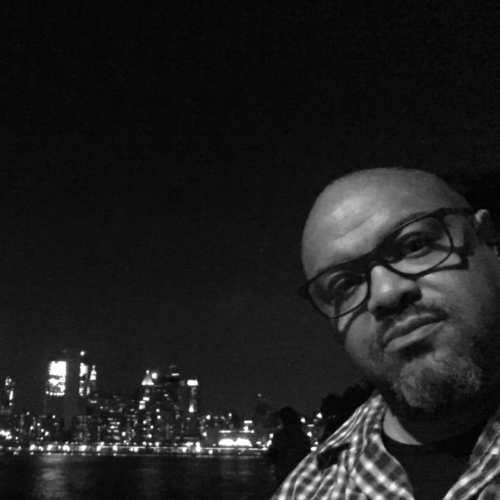 A black-and-white portrait of Michael Hobbs, standing against a dark background at night, looking at the camera while wearing glasses and a checkered shirt.