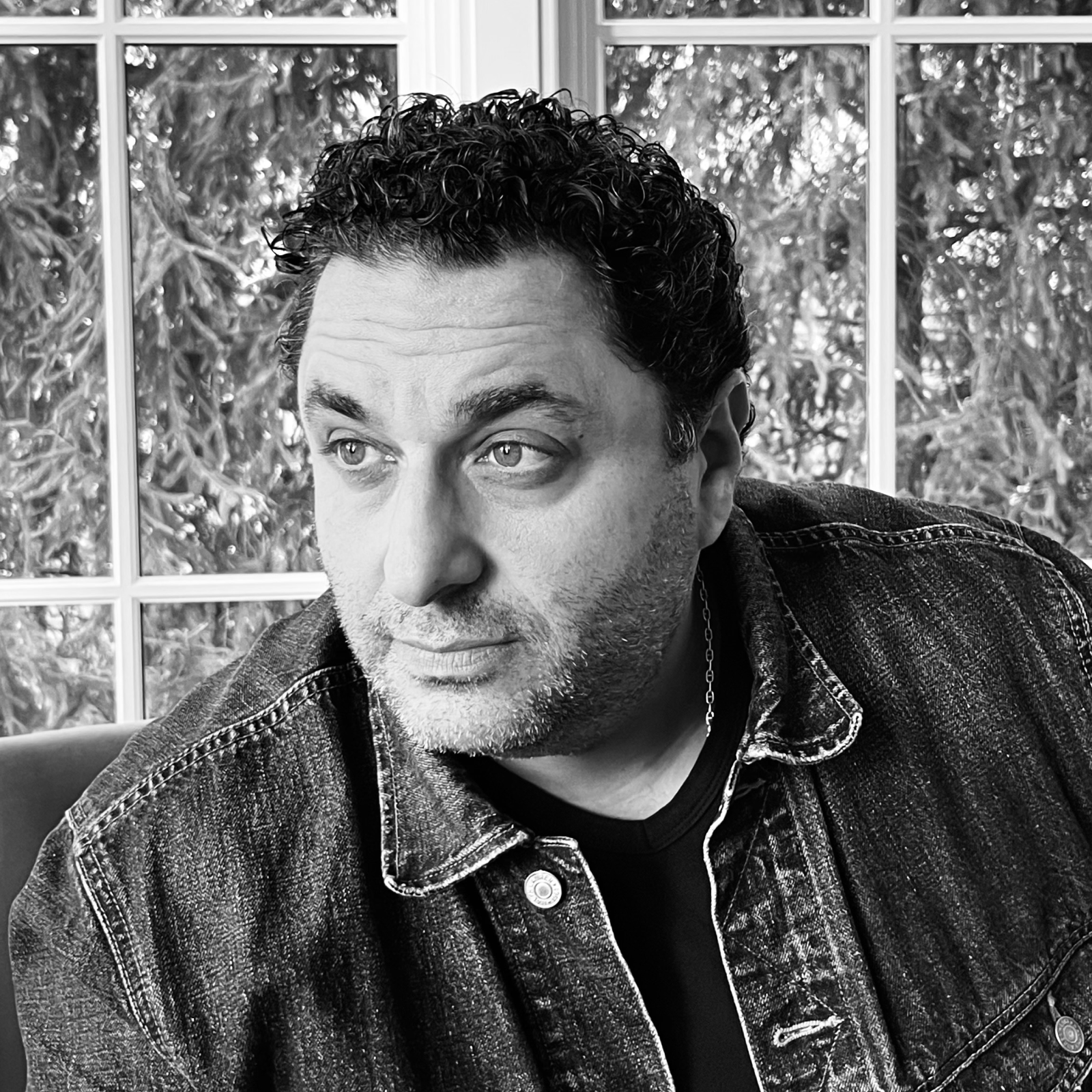 A black-and-white portrait of Michael Sarno, standing indoors, smiling slightly, wearing a dark jacket and glasses.