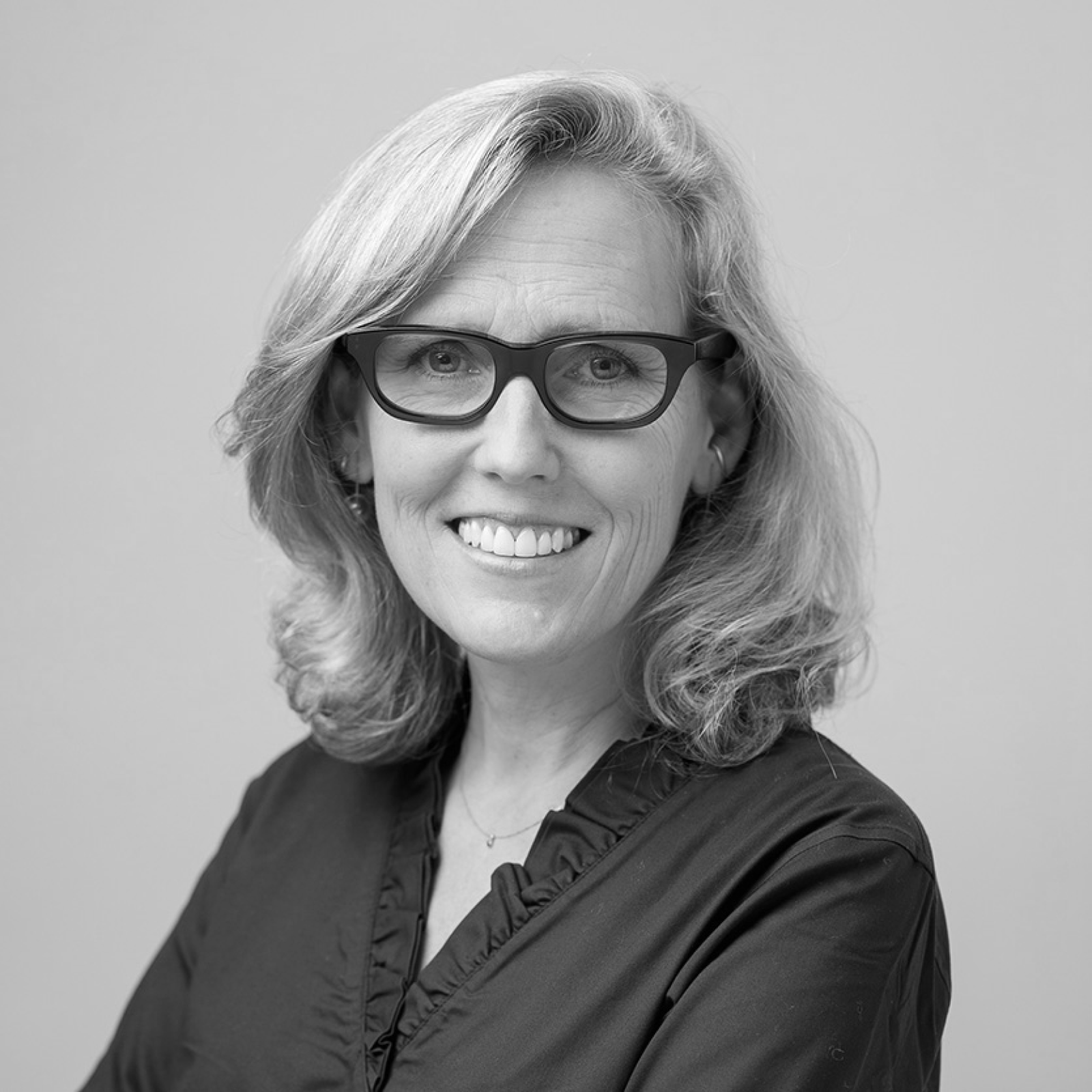 A grayscale portrait of a smiling woman with shoulder-length blonde hair and black-rimmed glasses. She is wearing a dark blouse and is looking directly at the camera.