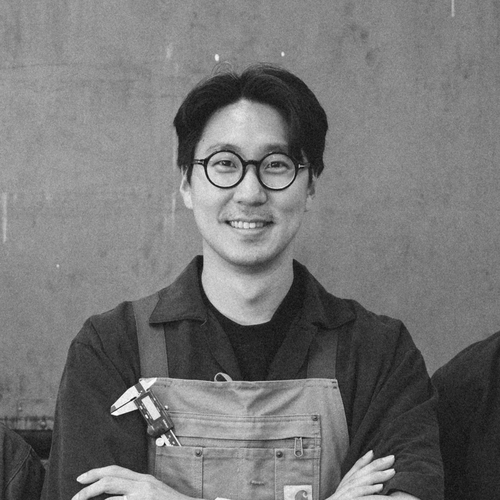 A black and white portrait of a man wearing round glasses and an apron, standing in front of a plain background and smiling.