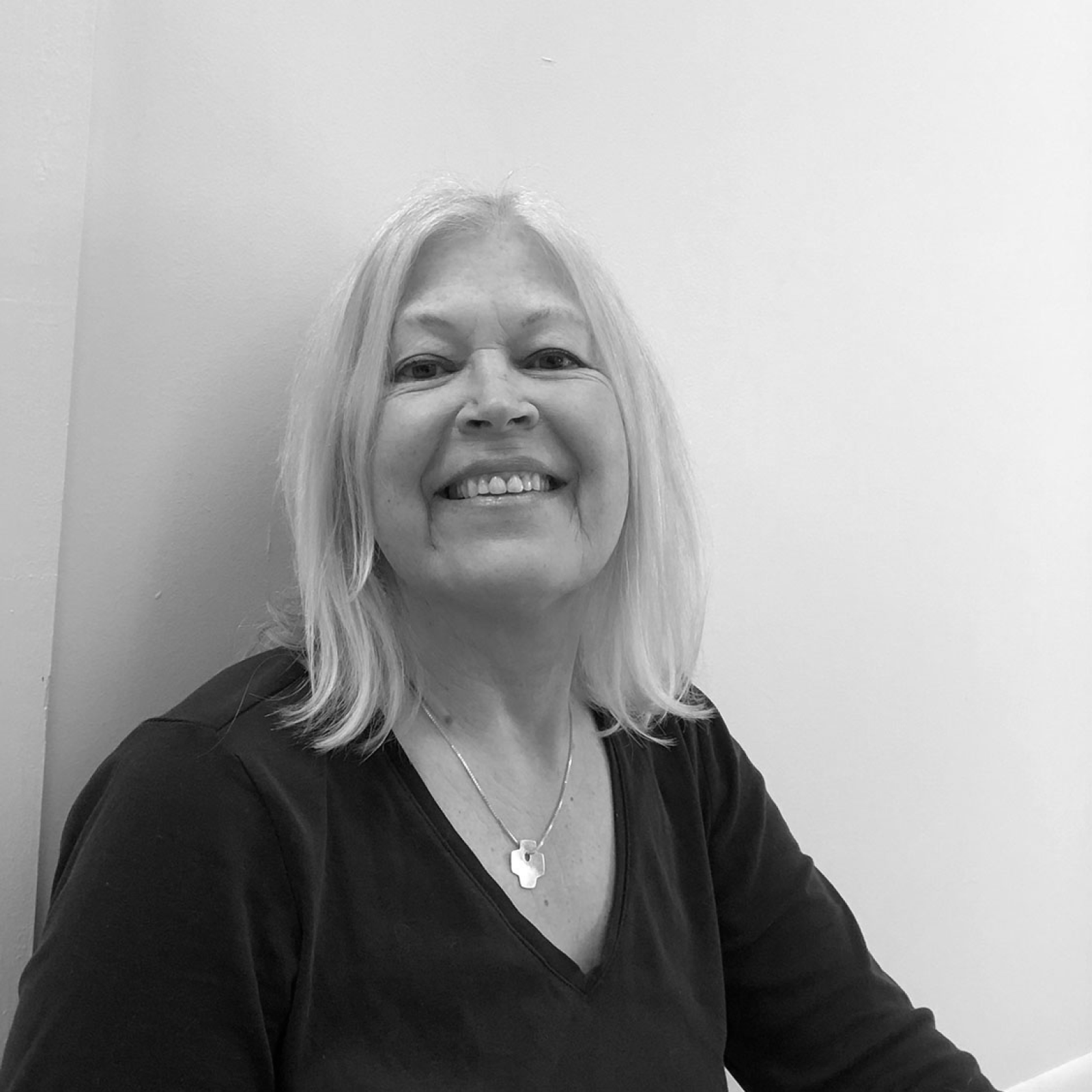 A grayscale portrait of a smiling woman with shoulder-length hair, sitting against a plain wall. She is wearing a dark shirt and a simple necklace, exuding a relaxed and friendly demeanor.