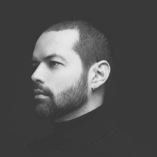 Rodolfo Agrella looks to the side, with a short beard and earring, wearing a black turtleneck against a dark background.