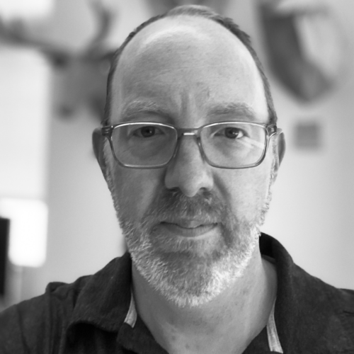 Steve Turbek faces the camera with glasses and a beard, wearing a dark polo shirt indoors with a soft background.