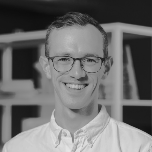 Tim Duschenes smiles while wearing glasses and a button-down shirt, with shelves in the background.