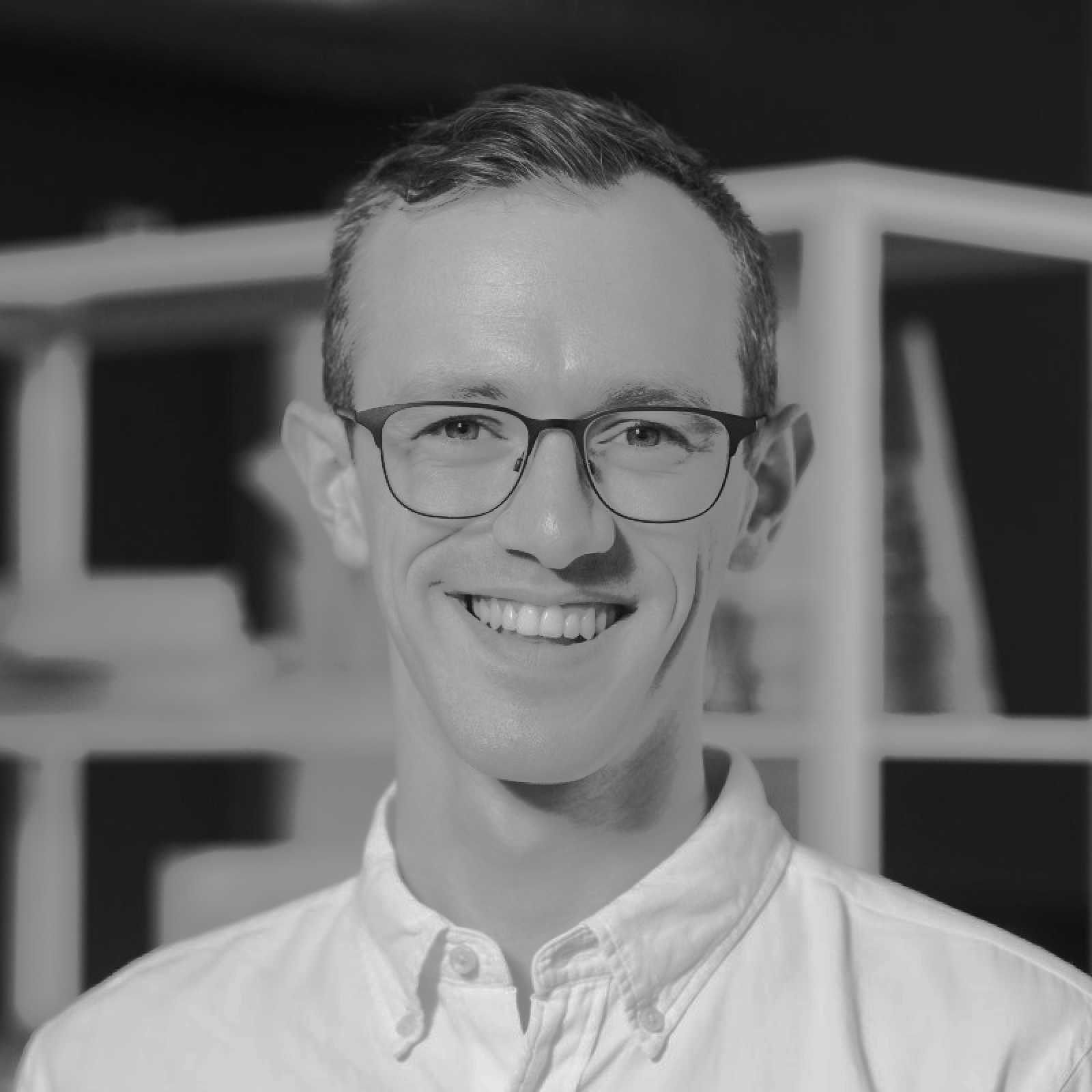 Tim Duschenes smiles while wearing glasses and a button-down shirt, with shelves in the background.