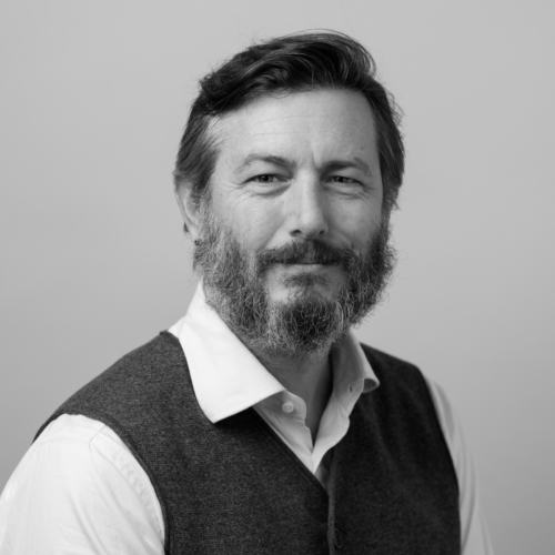 A black and white portrait of a man with short hair and a beard, wearing a white shirt and a dark vest, looking directly at the camera with a neutral expression.