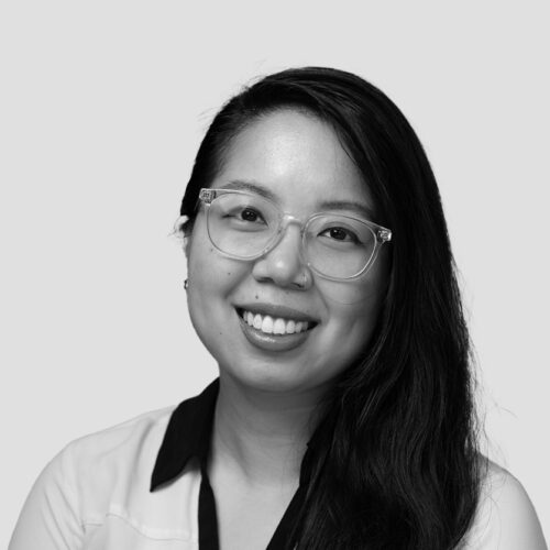 A black-and-white portrait of a young woman with long, dark hair, wearing clear-framed glasses and a white blouse with a black collar. She has a bright smile and looks directly at the camera. The background is a plain, light gray color.