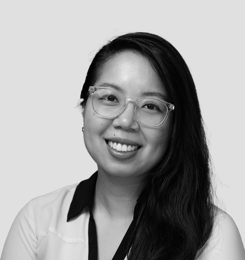 A black-and-white portrait of a young woman with long, dark hair, wearing clear-framed glasses and a white blouse with a black collar. She has a bright smile and looks directly at the camera. The background is a plain, light gray color.