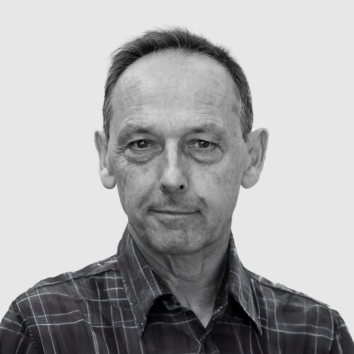 A black-and-white portrait of a middle-aged man with short hair, wearing a plaid button-up shirt. He has a neutral expression with a hint of a smile and gazes directly at the camera. The background is a plain, light gray color.