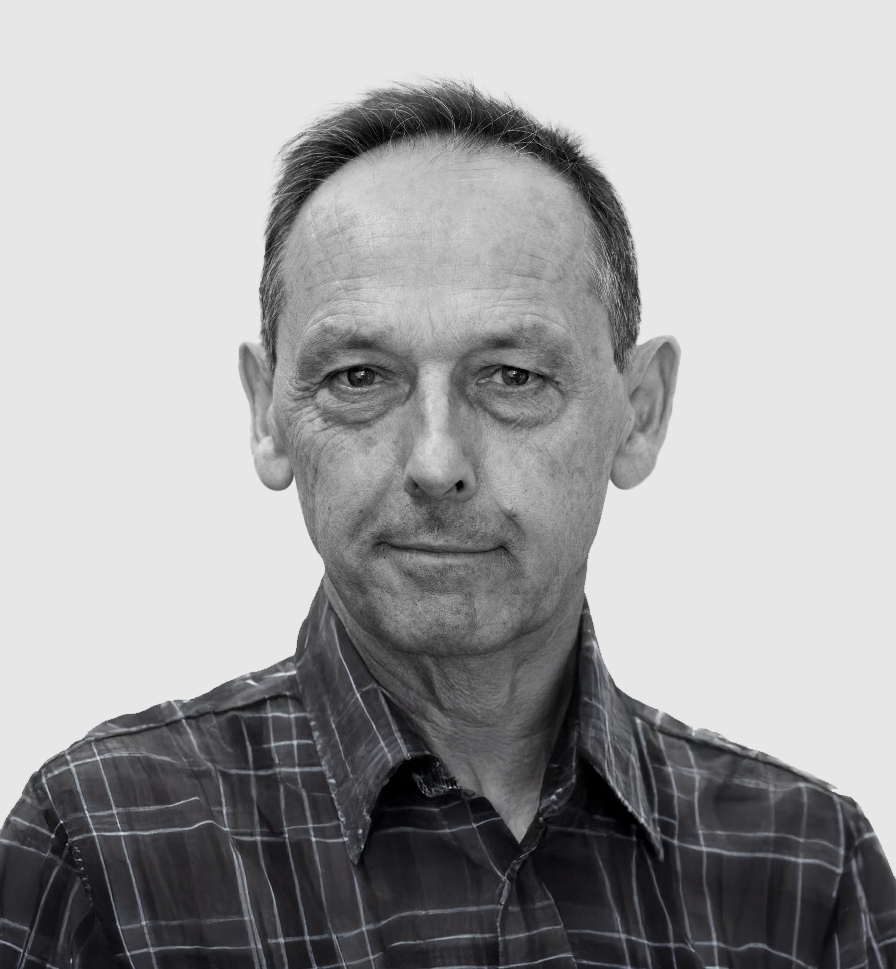 A black-and-white portrait of a middle-aged man with short hair, wearing a plaid button-up shirt. He has a neutral expression with a hint of a smile and gazes directly at the camera. The background is a plain, light gray color.