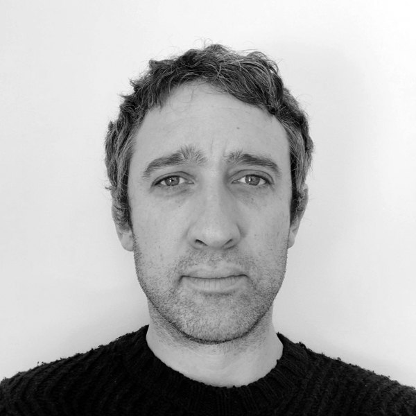 A black-and-white close-up portrait of a man with dark hair and light stubble. He is wearing a dark sweater and looking directly at the camera with a neutral expression. The background is plain white.