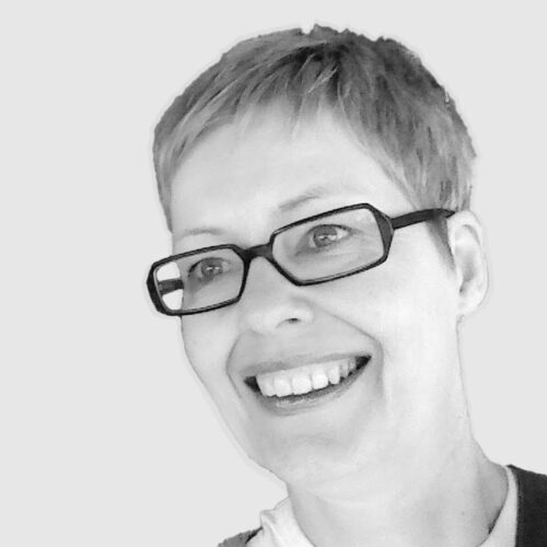 A black-and-white portrait of a woman with short, light-colored hair, wearing rectangular glasses and a sweater over a white shirt. She has a warm smile and is looking slightly to the side. The background is a plain, light gray color.