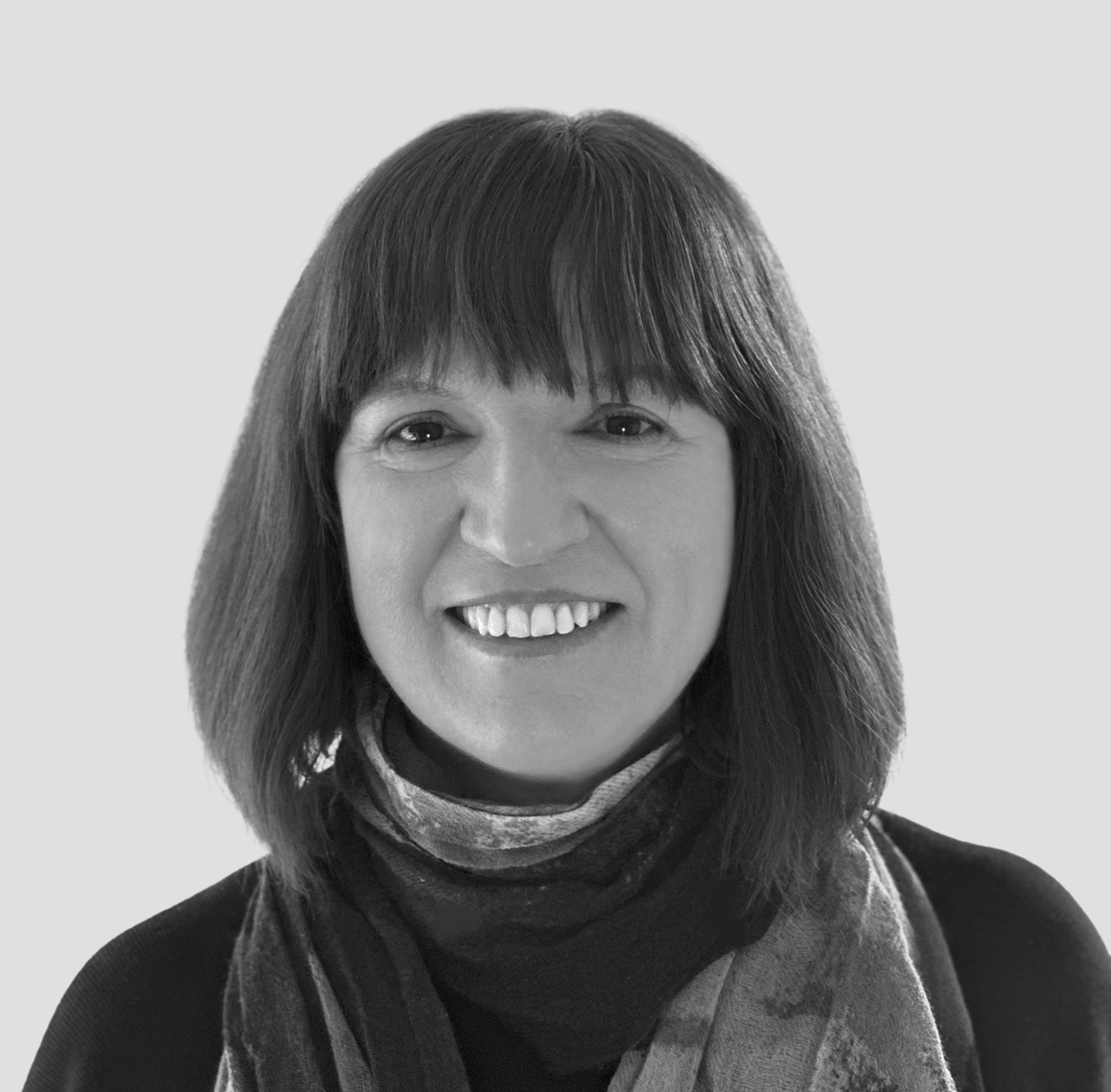 A black-and-white portrait of a woman with shoulder-length dark hair and bangs. She is wearing a dark top with a patterned scarf wrapped around her neck. She has a warm smile and looks directly at the camera. The background is a plain, light gray color.