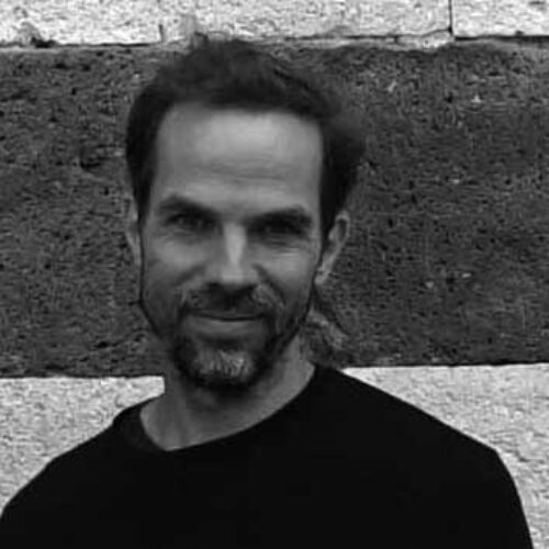 A black-and-white portrait of a man with short dark hair and a beard. He is wearing a dark long-sleeved shirt and is smiling slightly. The background consists of a stone or brick wall with a textured pattern.