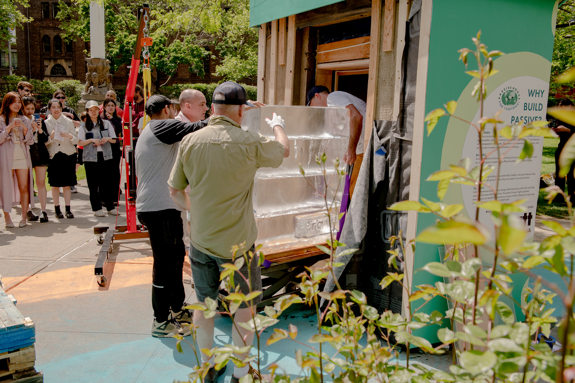 Four people move large stack of ice blocks out of a small structure as students look on