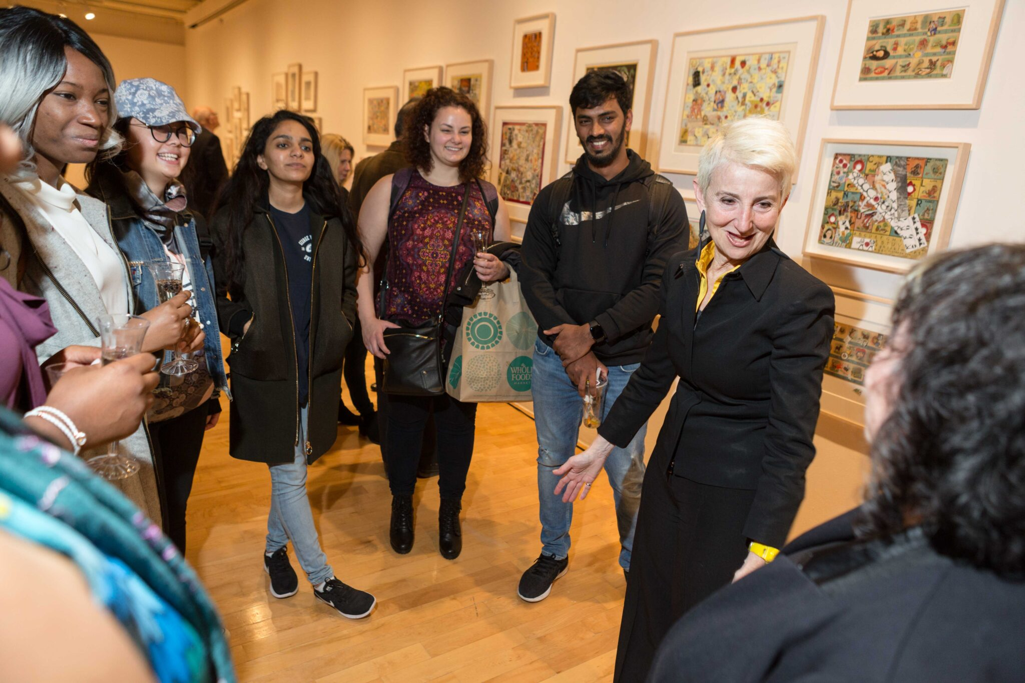 A group of people stand in an art gallery, engaged in conversation. The group consists of diverse individuals, including Pratt President Frances Bronet leading the discussion. The gallery walls are adorned with framed artworks featuring colorful illustrations. Some people in the group are holding drinks, and one person carries a tote bag. The atmosphere is lively and interactive, with everyone appearing interested and engaged in the conversation.