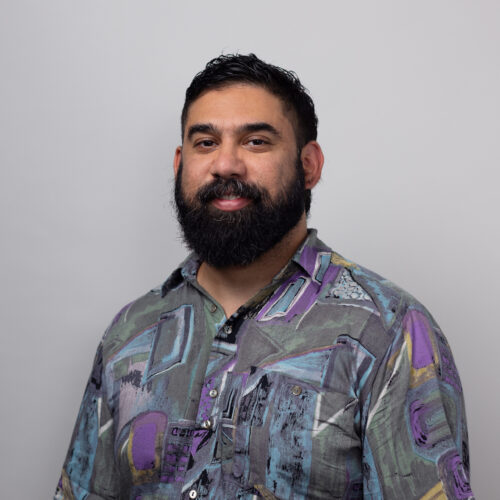 Portrait of a man with a full beard, wearing a multicolored abstract patterned shirt, looking at the camera with a slight smile.