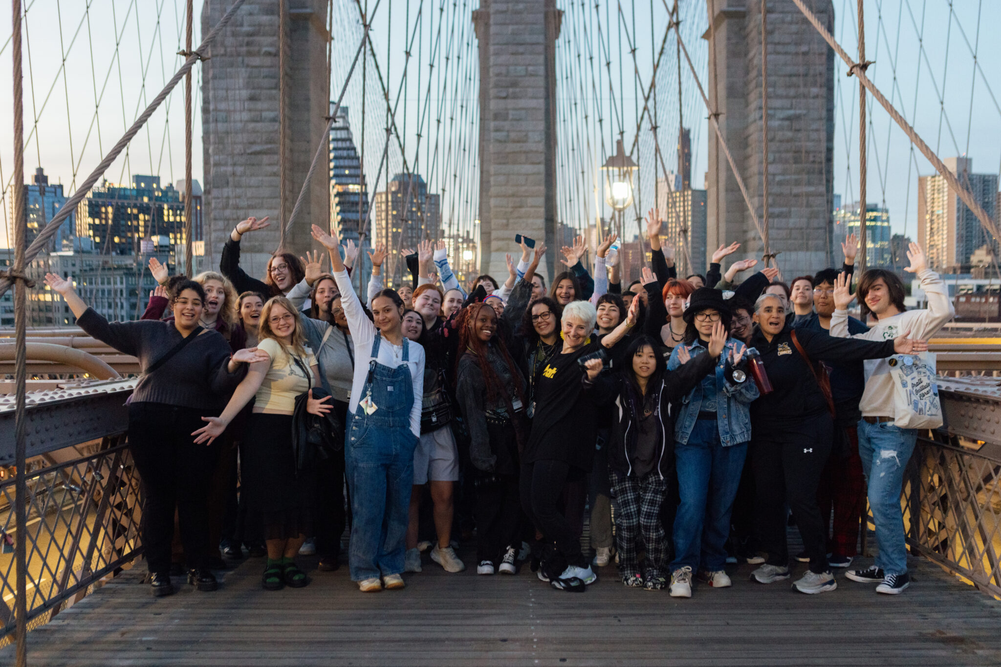 A large group of people, predominantly students, gathered on the Brooklyn Bridge, posing for a group photo. They are smiling and waving their hands in the air, creating a lively and celebratory atmosphere. The city skyline is visible in the background as dusk or dawn light casts a warm glow, adding to the scenic and joyful moment captured during the group outing.