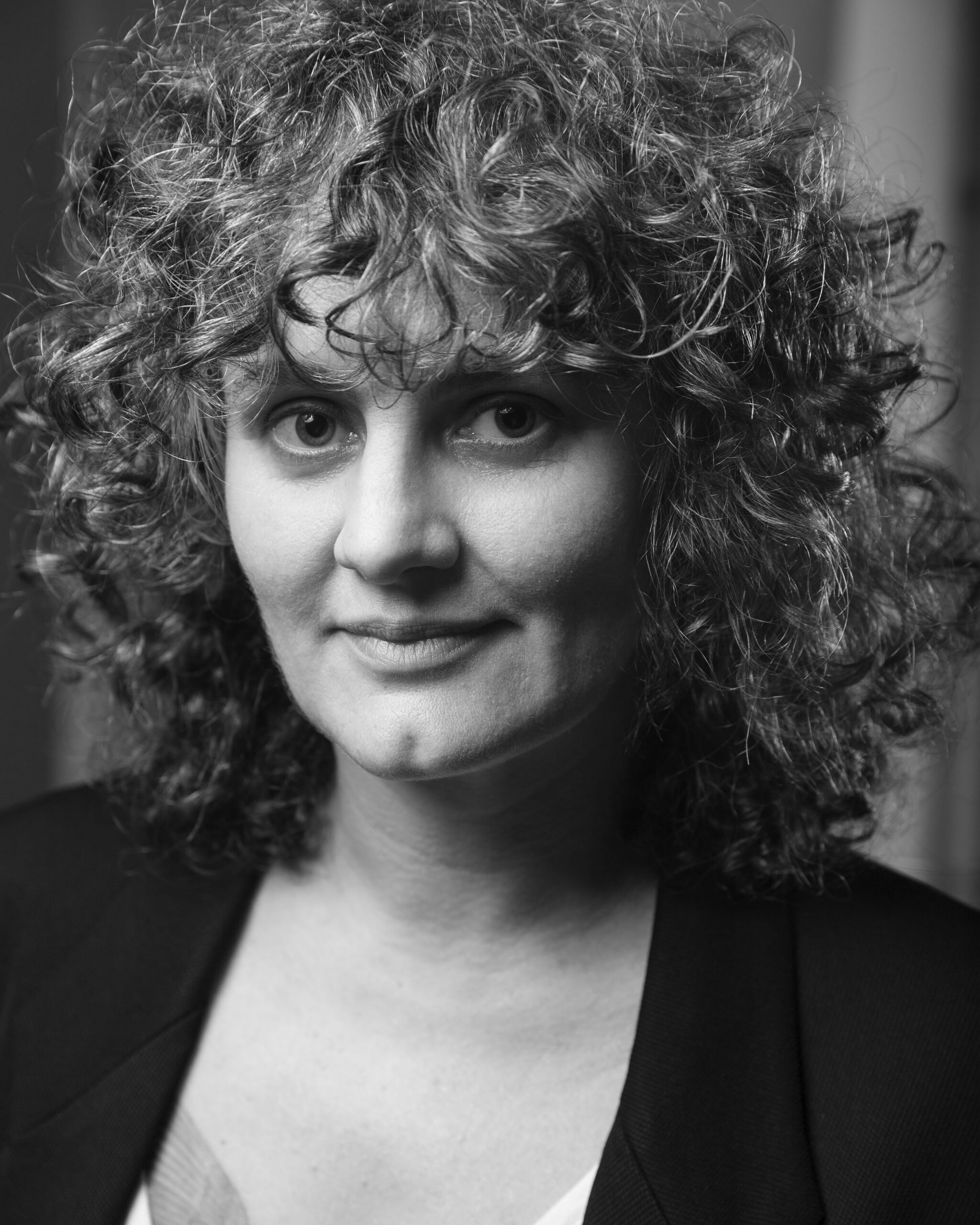 Black and white portrait of a woman with voluminous curly hair, wearing a dark blazer over a light top, looking at the camera with a slight smile and intense eyes.