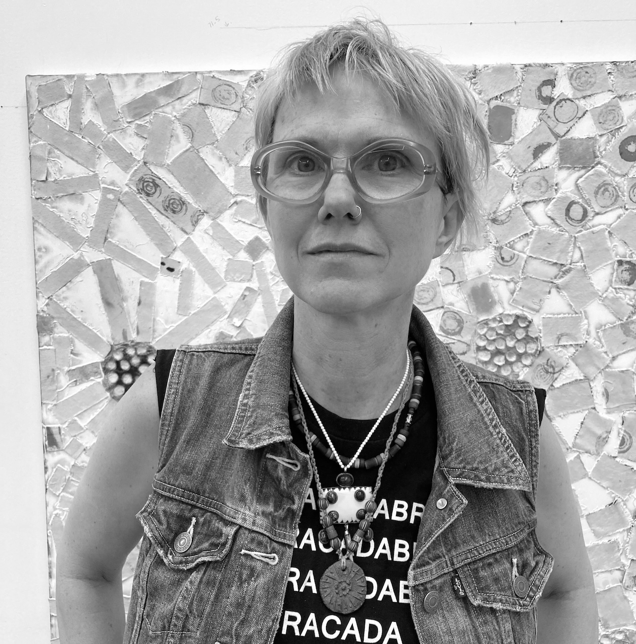 Black and white portrait of a woman with short light hair and round glasses, standing in front of an abstract mosaic artwork, wearing a denim vest and various necklaces.