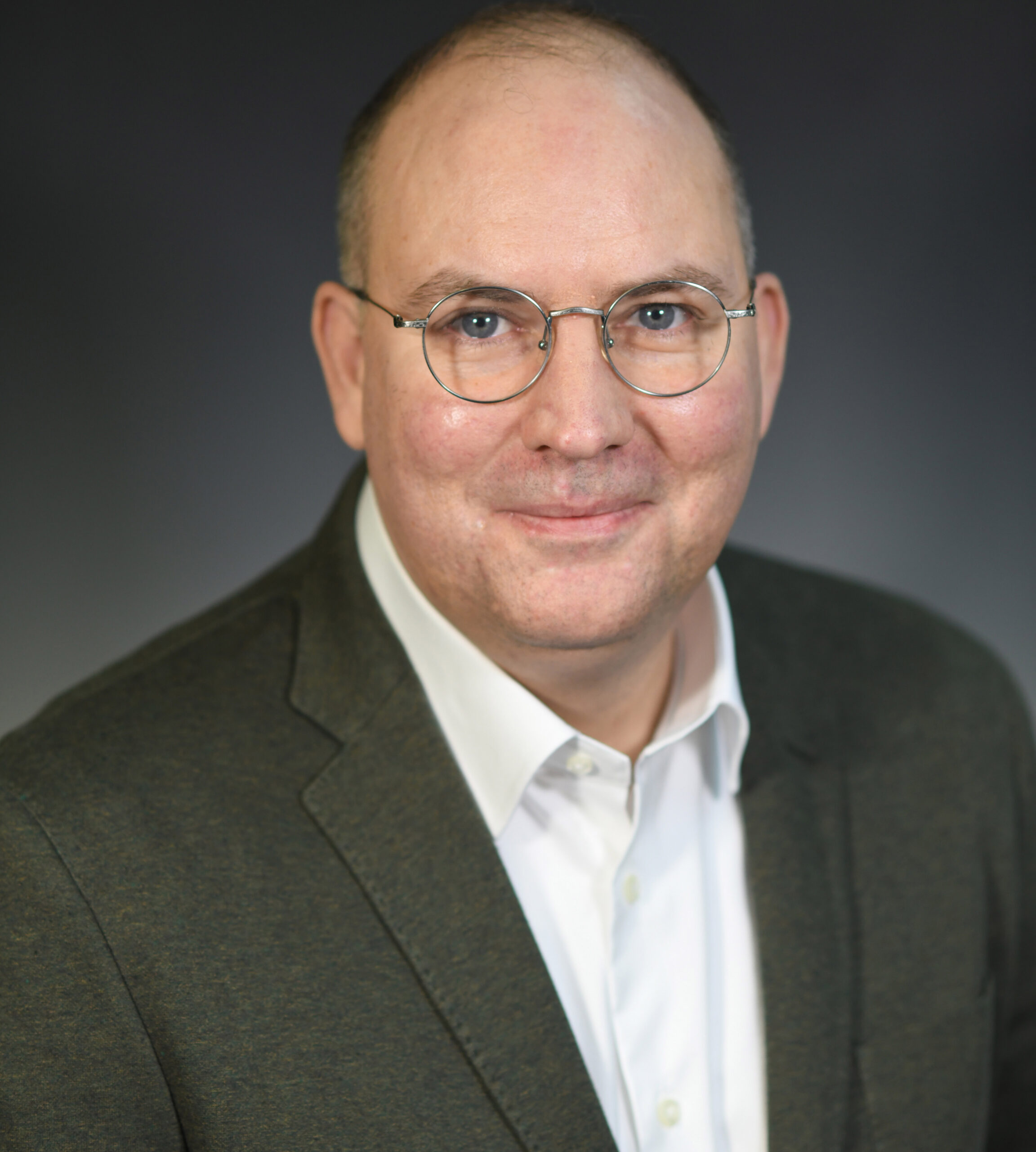 Portrait of a middle-aged man wearing round wire-framed glasses and a dark green blazer over a white shirt, smiling slightly, with a neutral gray background.