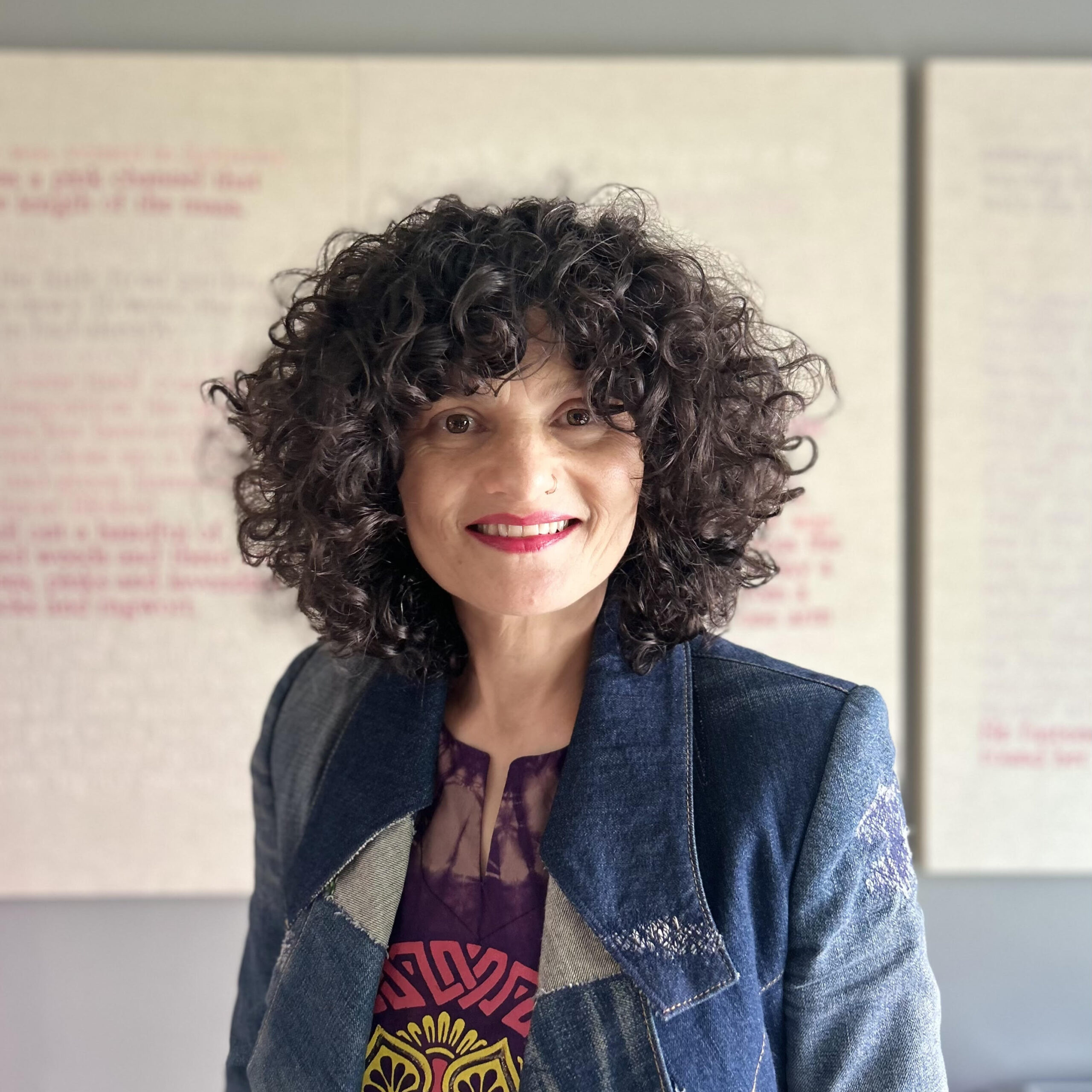 Portrait of a woman with voluminous curly black hair, wearing a denim jacket and a colorful graphic t-shirt, standing against a text-covered wall, smiling brightly.