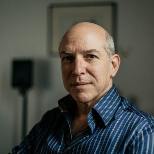 Horizontal portrait of a middle-aged man with a bald head, wearing a striped blue shirt, looking contemplative as he gazes slightly off-camera in an indoor setting.