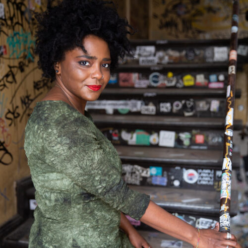 Portrait of a woman with curly black hair and bright red lipstick, standing in a graffiti-covered alley, wearing a textured green dress and looking back over her shoulder with a confident smile.