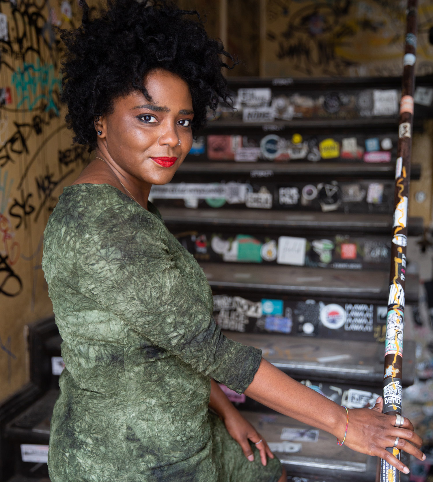 Portrait of a woman with curly black hair and bright red lipstick, standing in a graffiti-covered alley, wearing a textured green dress and looking back over her shoulder with a confident smile.