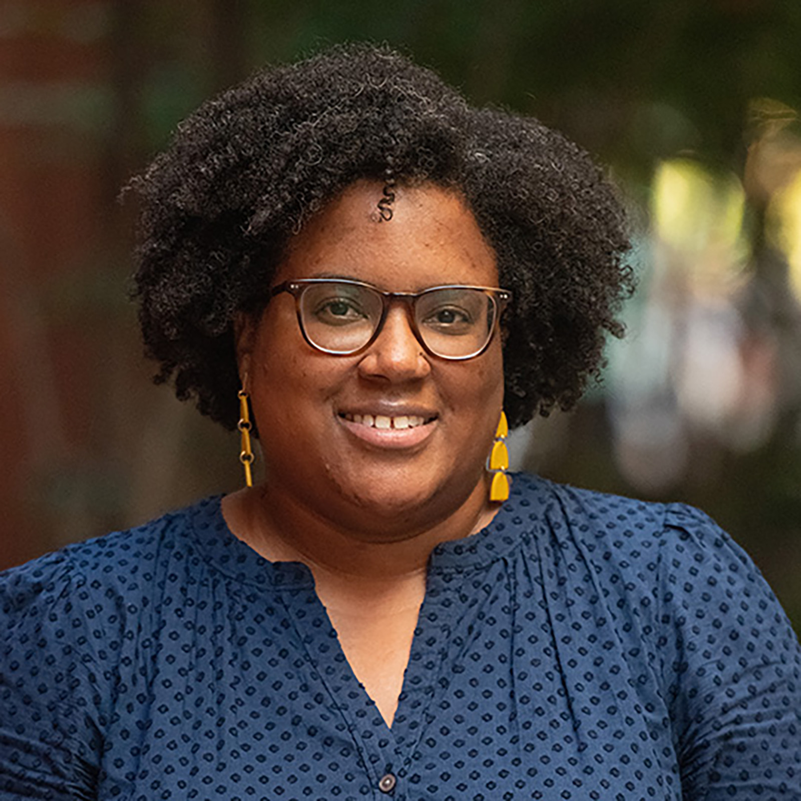 A close-up portrait of a woman with natural hair styled in tight curls, wearing glasses with dark frames. She is smiling confidently and is dressed in a navy blue top with a subtle polka-dot pattern. Yellow earrings add a pop of color, and the blurred green foliage in the background contributes to a warm, inviting tone in the image.