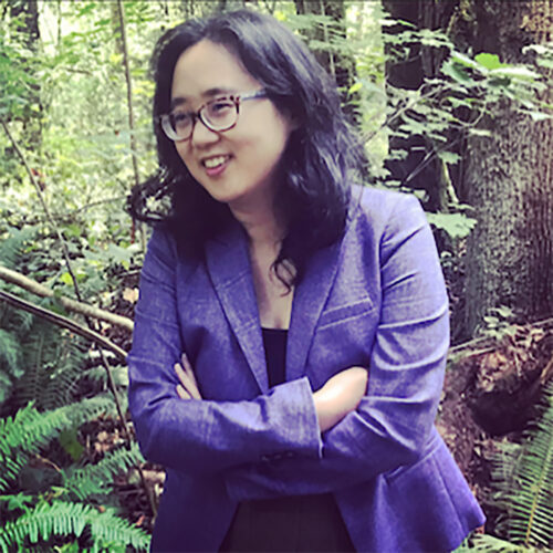 This is a photo of a woman standing in a natural forest setting, arms crossed and smiling softly. She has shoulder-length black hair, wears glasses, and is dressed in a deep purple blazer. The lush greenery and trees in the background give the image a peaceful, contemplative atmosphere.