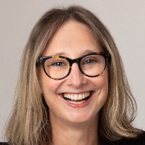 This is a professional headshot of an individual with light brown, shoulder-length hair wearing glasses with dark frames. The subject is smiling warmly and is dressed in professional attire, with a black blazer and light-colored shirt. The background is neutral, and the lighting is soft, creating a polished and approachable look.