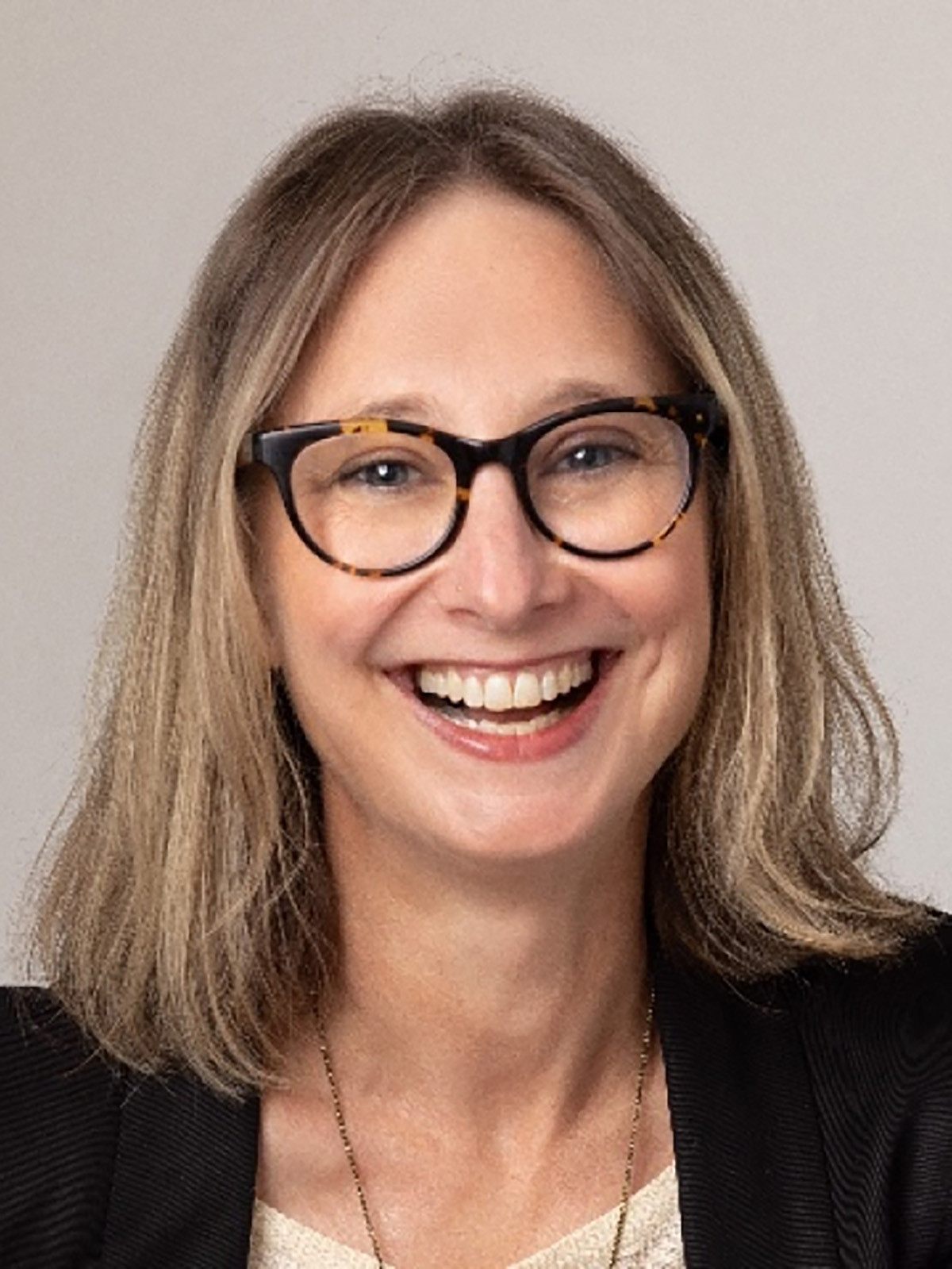 This is a professional headshot of an individual with light brown, shoulder-length hair wearing glasses with dark frames. The subject is smiling warmly and is dressed in professional attire, with a black blazer and light-colored shirt. The background is neutral, and the lighting is soft, creating a polished and approachable look.