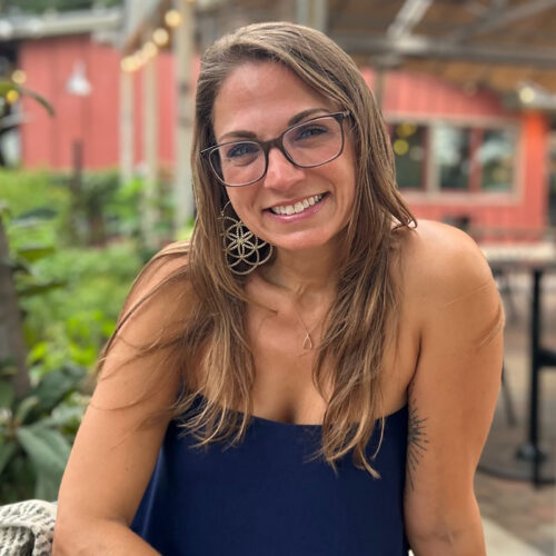 This image portrays a woman seated outdoors, smiling brightly at the camera. She has long light brown hair, wears glasses, and is dressed in a sleeveless navy blue top. Her earrings are large and geometric in design, and the background shows a blurred patio area with plants and outdoor seating, suggesting a relaxed atmosphere.