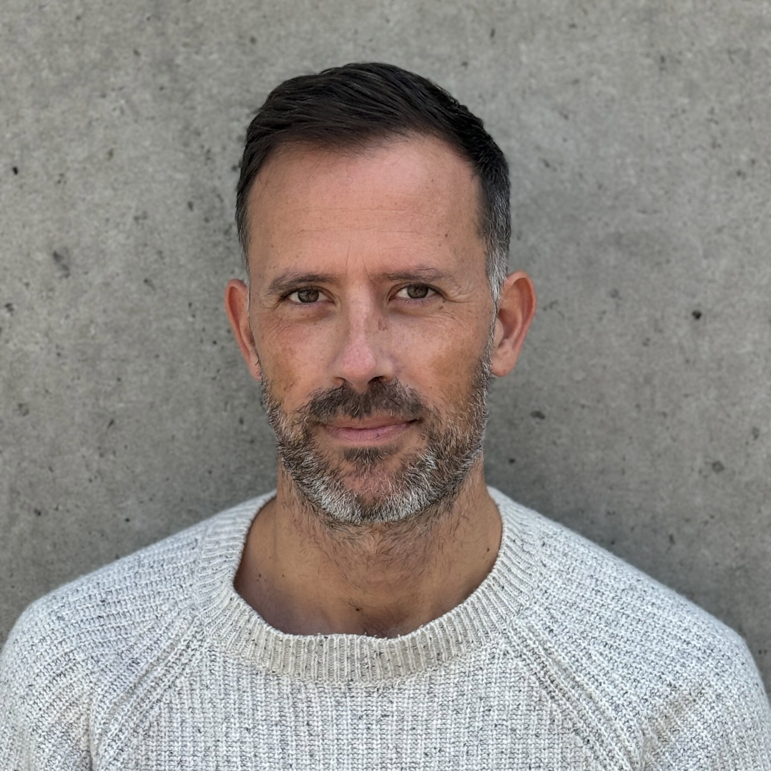 Portrait of a middle-aged man with short, light brown hair and stubble, wearing a white sweater, standing against a textured gray background, looking directly at the camera with a serious expression.