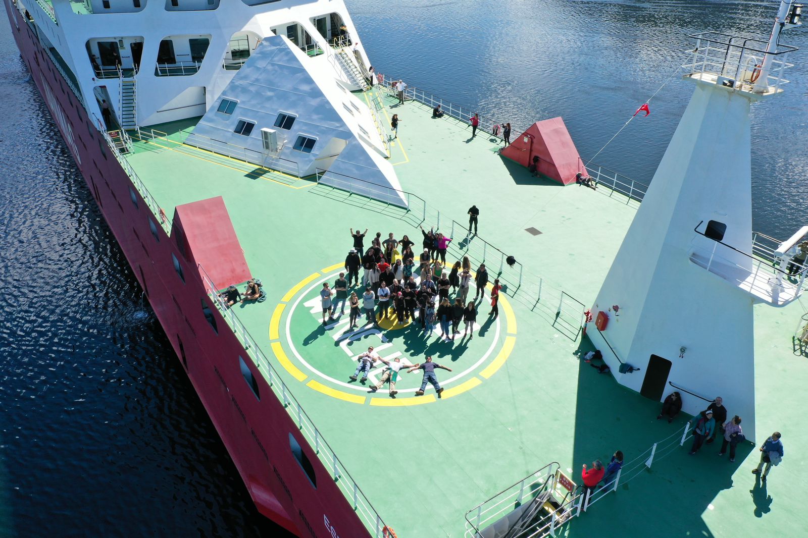 This aerial image captures a large group of people on the helipad of a ferry. The ferry, painted in vibrant red and white, floats on calm blue waters. The deck is marked with a large yellow and green helicopter landing circle. People are seen engaging in various activities; some are walking, others standing and chatting, and a few are participating in what appears to be a group dance or exercise. The environment is bright and clear, emphasizing the lively atmosphere of the gathering.